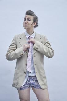 © Jana Rothe Der Drag King-Charakter Richard P. Dick Van Johnson trägt eine beige Anzugjacke, keine Hose und eine passende rosa Krawatte und Unterwäsche. Er repariert seine Krawatte.