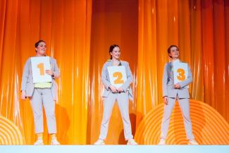 Drei Performerinnen in grauen Anzügen stehen vor einem orangefarbenen Vorhang und halten drei Tafeln mit den Nummer 1, 2 und 3 in den Händen.