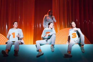 Drei Performerinnen in grauen Anzügen sitzen auf einem Bühnenrand und halten drei Tafeln mit den Nummer 1, 2 und 3 in den Händen. Im Hintergrund steht eine Frau vor einem orangefarbenen Vorhang und spricht in ein Mikro. 