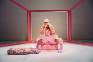 A performer, dressed in a loose sleeveless jersey, pink sports pants and a white bandana, holds a cell phone to his ear with his right hand. He is sitting on a pink cushion. In front of him in the left half of the image is a pink plush garment on the floor. His playing surface is framed by a framework of pink struts. 