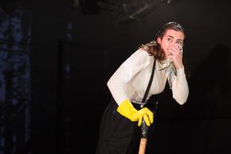© Michael Felsch Zu sehen: Die Schauspielerin Marie Philipp in der Rolle des Kaufmannes Herr Michael Fischer. Sie trägt einen weißen Rollkragenpullover, eine schwarze Hose mit Hosenträgern und einen gelben Handschuh. In der rechten Hand hält sie ein Stock, in der linken ein Taschentuch. Sie guckt ängstlich zurück, während sie das Taschentuchan ihre Nase hält.
