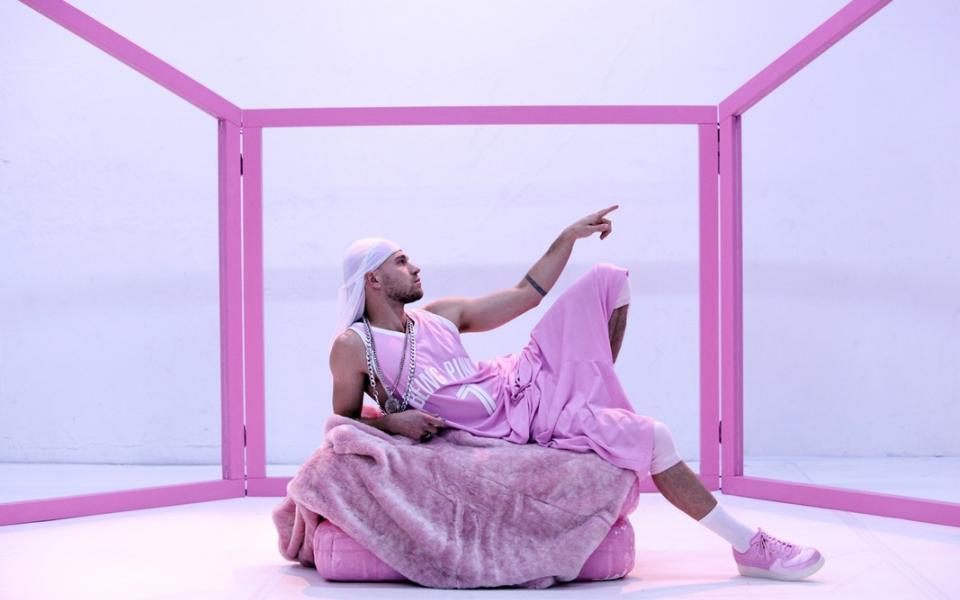 A performer, dressed in a loose sleeveless jersey, pink sports pants and a white bandana, lies on a pink cushion. His left index finger points into the distance, and his left leg is bent. The pose is modeled after Michelangelo's ceiling fresco, "The Creation of Adam." In the background, the stage set is framed by a framework of pink struts. 