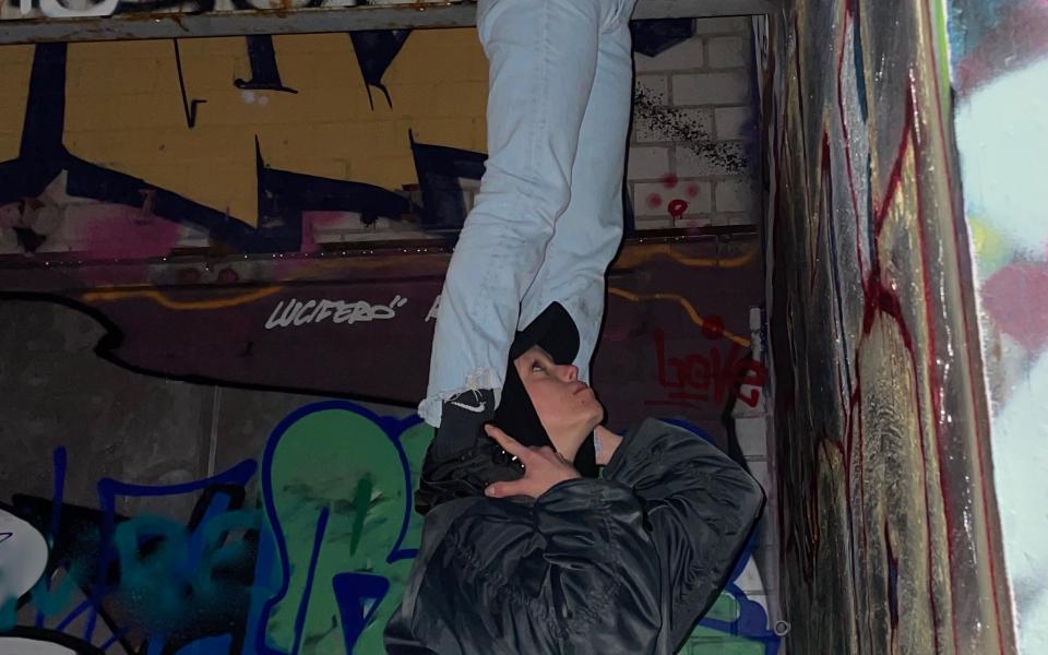 In front of a wall with graffiti, a person stands on the shoulders of another person and reaches up to the ceiling. The person below is also looking up. 