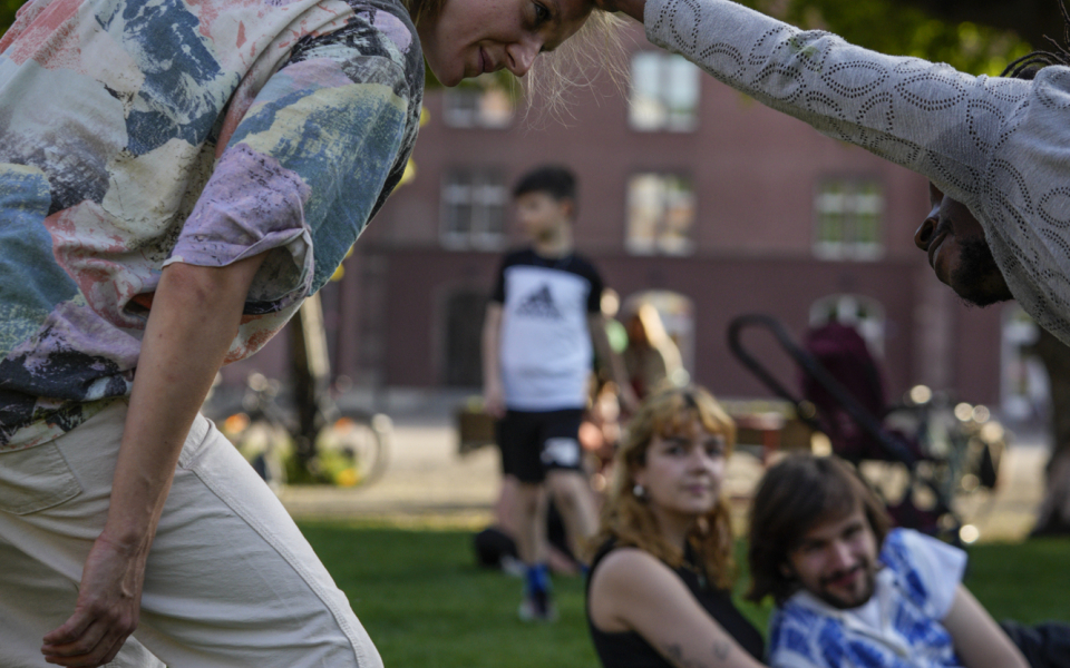 Melissa is a white woman with long blonde hair tied back in a ponytail. She is wearing a colourful shirt and white trousers. She smiles as she bends her knees and tilts her head and face forward towards another dancer who smiles at her and holds her head with his outstretched right hand. They are in a park with green grass and a green lush tree in the background and an audience watching from close by.