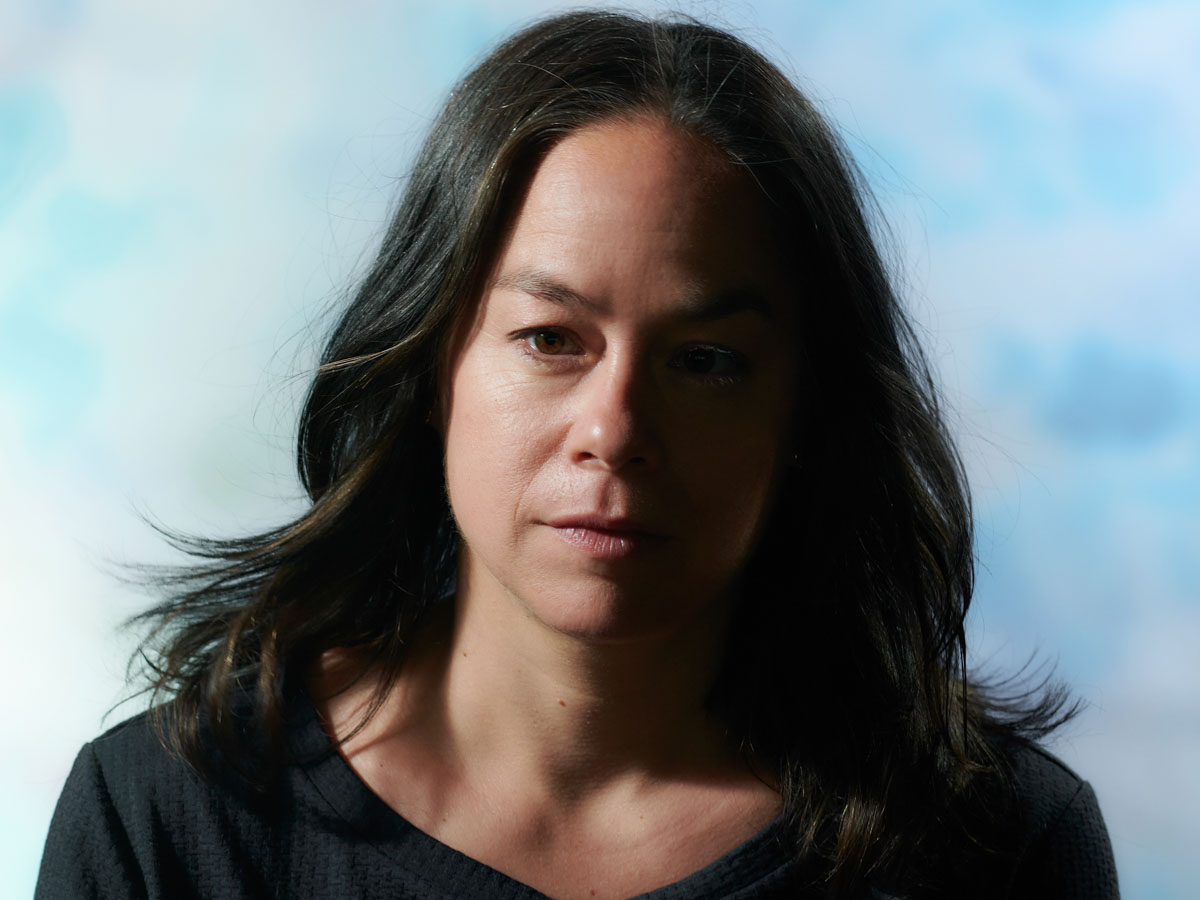 Portrait photo of Mirjam Gurtner against a light blue cloudy background. A white woman, with brown eyes and brown long hair worn loose, and a dark blue T-shirt.