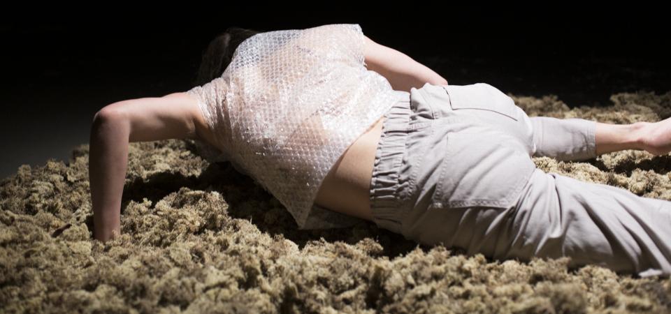 A person lies on a light surface of organic material - moss. The person is turned away from, props himself up a bit with his hands and keeps his head lowered. The person wears light pants and a short-sleeved shirt made of bubble wrap. The background is black. 