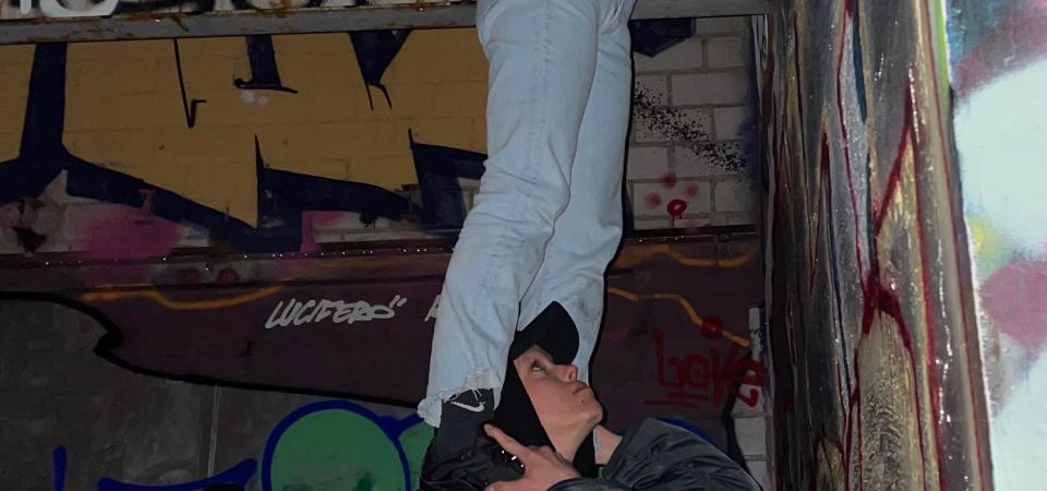 In front of a wall with graffiti, a person stands on the shoulders of another person and reaches up to the ceiling. The person below is also looking up.  